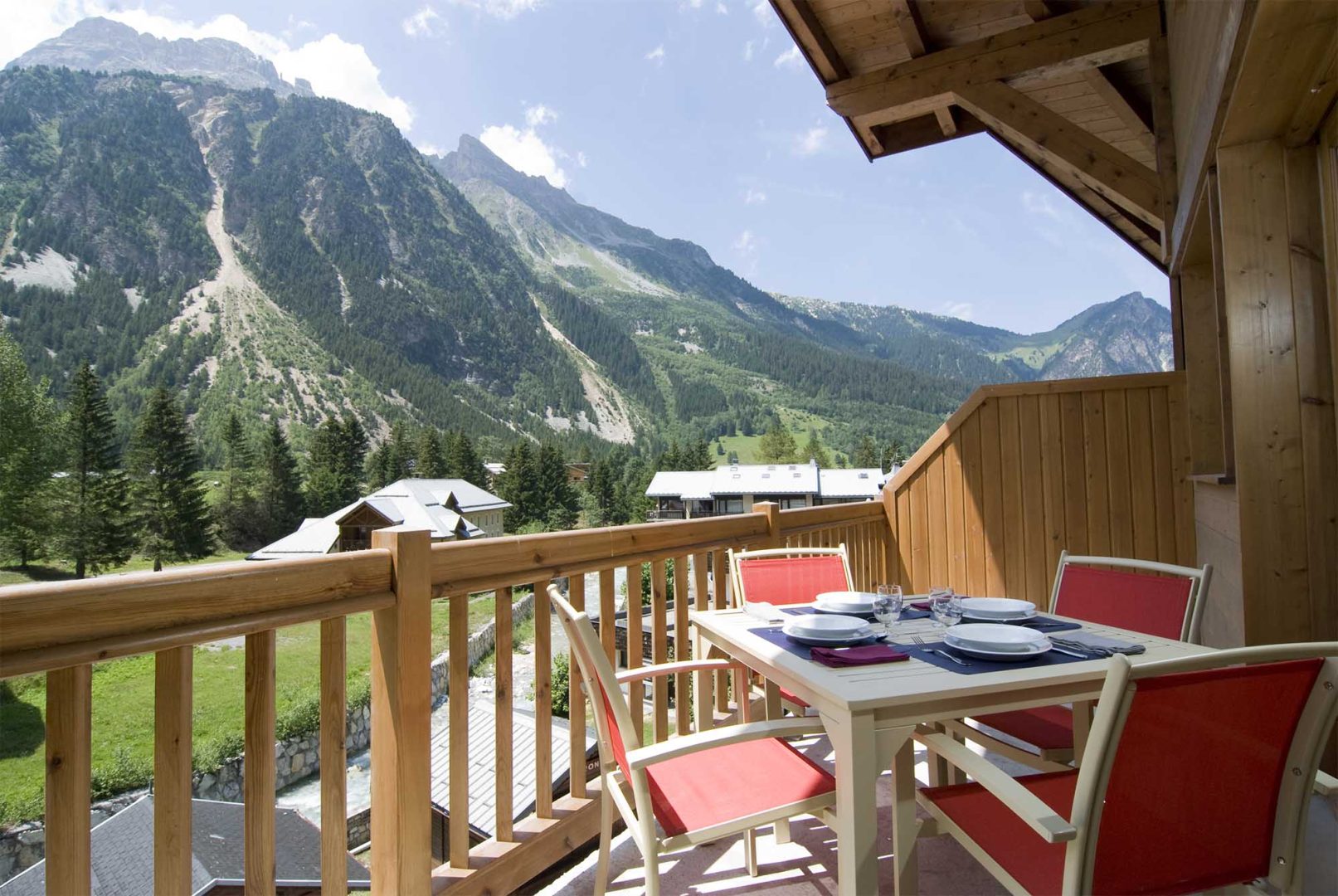 Image de la résidence Les Jardins de la Vanoise de Madame Vacances : balcon en bois avec une table et quatre chaises, offrant une vue sur des montagnes verdoyantes et des chalets alpins sous un ciel ensoleillé