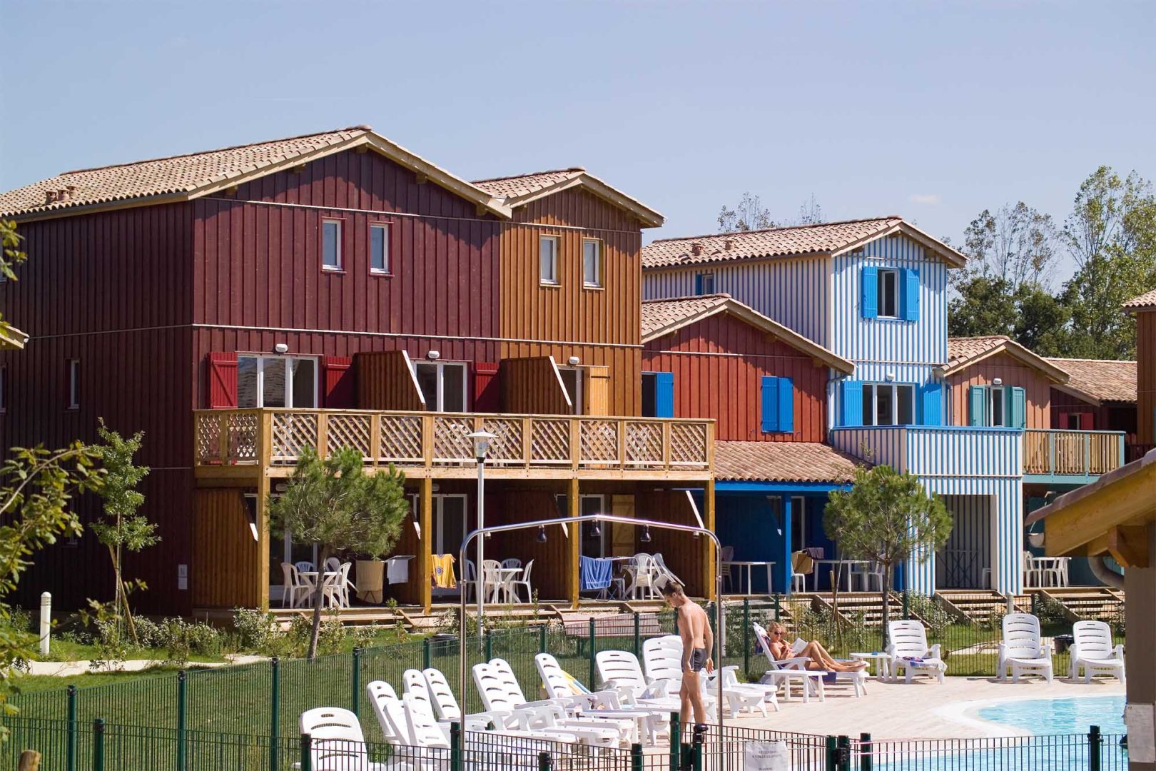 Image de la résidence Les Rives Marines de Madame Vacances : bâtiments en bois avec balcons, vues depuis une piscine avec des transats et des vacanciers au premier plan